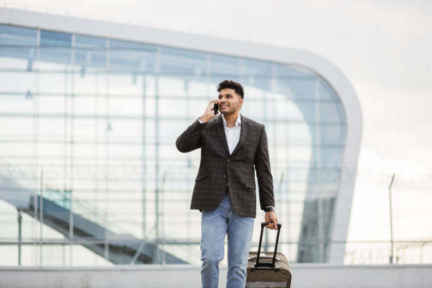 istockphoto-1384210736-612×612 Portrait of young handsome Indian business man walking outside to station or airport talking on the phone and carrying suitcase.